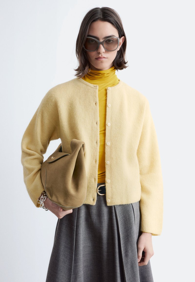 Junge Frau mit großen Sonnenbrillen, einer gelben Strickjacke über einem senffarbenen Rollkragenpullover, grauem Rock, die eine hellbraune Wildlederhandtasche hält.