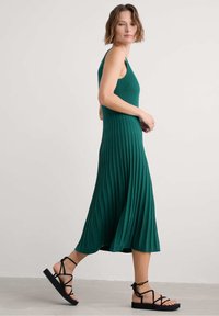 Green ribbed midi dress with a fitted bodice, flared skirt, and thin shoulder straps, paired with black strappy sandals.