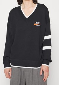 Person wearing black V-neck Nike sportswear sweater with white trim and two white stripes on the right sleeve, paired with gray pants.