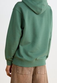 Zelená mikina s volným střihem, vyrobená z měkké tkaniny. Má kapuci se stahovací šňůrkou a žebrované manžety. Kombinováno se světle hnědými kalhotami.