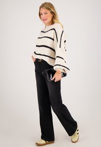 Vrouw die een witte trui met zwarte strepen draagt, zwarte broek, beige sneakers en een zwarte clutch vasthoudt, staand tegen een effen achtergrond.