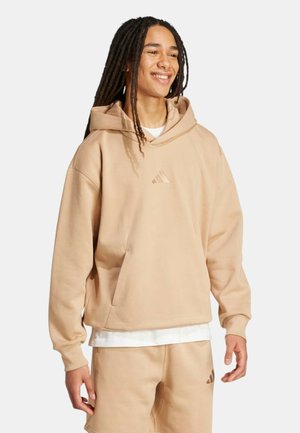 Giovane con lunghi dreadlocks sorridente, indossa una felpa con cappuccio beige e pantaloncini abbinati con dettagli di logo sottili su sfondo chiaro.