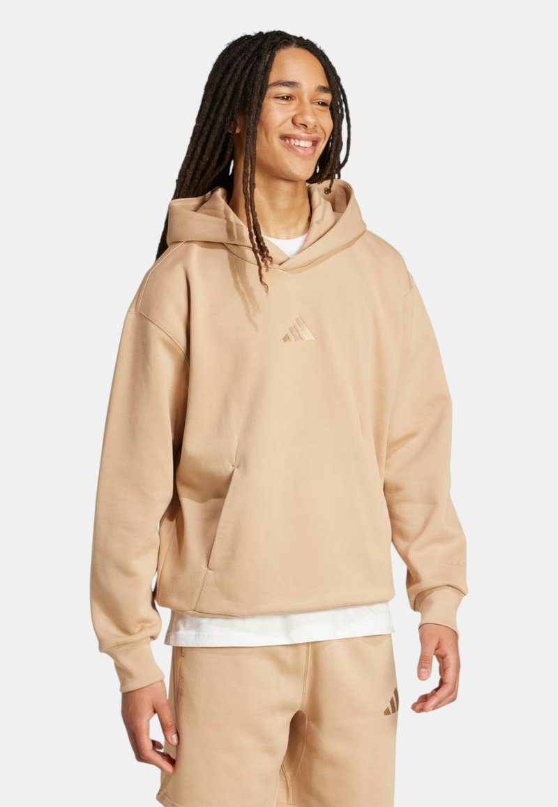 Giovane con lunghi dreadlocks sorridente, indossa una felpa con cappuccio beige e pantaloncini abbinati con dettagli di logo sottili su sfondo chiaro.