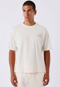 Korte mouw crèmekleurig T-shirt van zacht materiaal, met een subtiele logo in beige op de linkerkant van de borst. Relaxte pasvorm met een ronde hals.