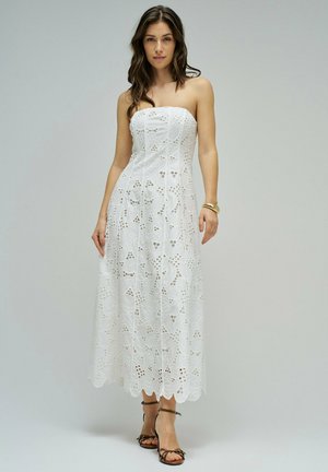 Femme portant une robe midi blanche en dentelle œillet sans bretelles et des talons noirs à brides, debout devant un fond gris clair uni.