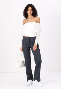 White off-the-shoulder top with ruched texture, paired with dark striped trousers. Model holds a white handbag and wears white sneakers.