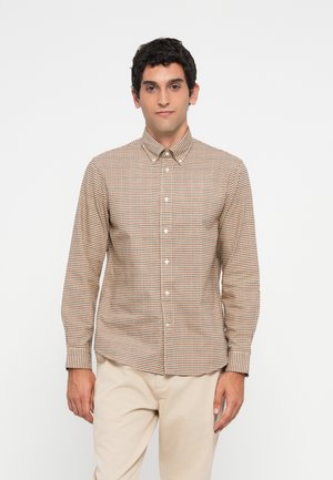 Man wearing beige and brown checkered button-up shirt and light beige pants standing against a white background.