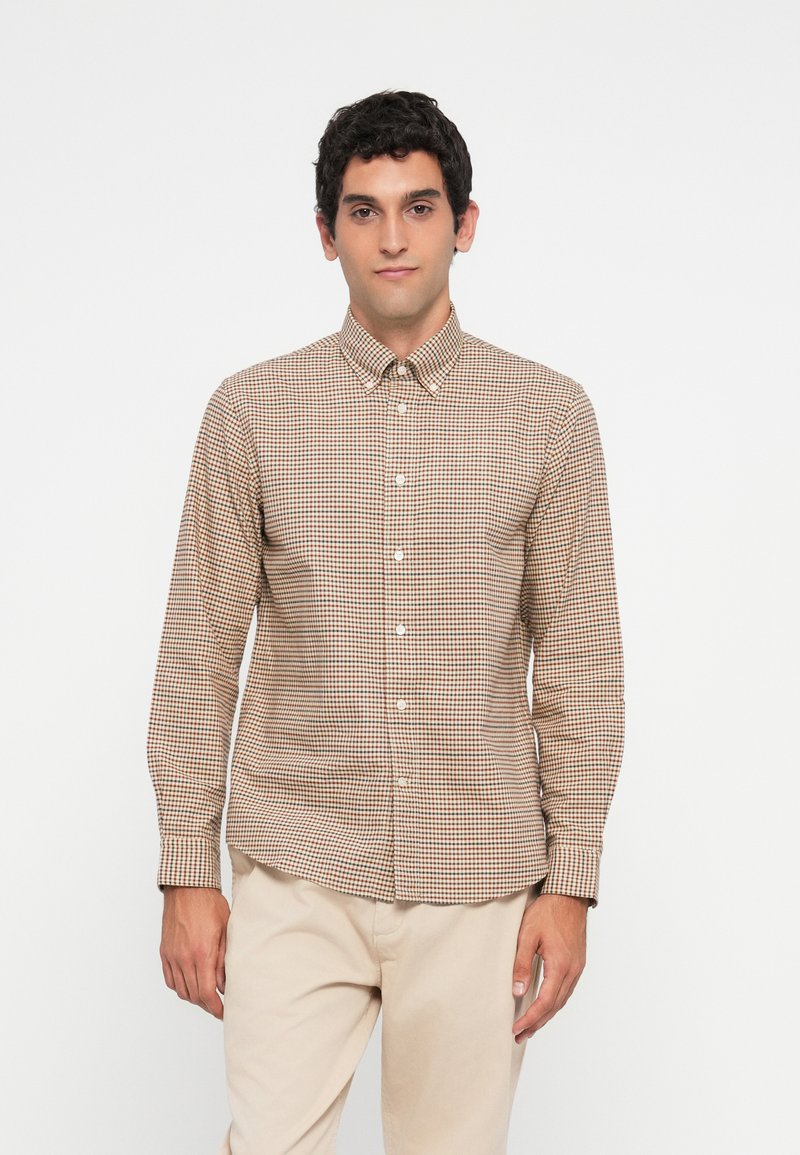 Man wearing beige and brown checkered button-up shirt and light beige pants standing against a white background.