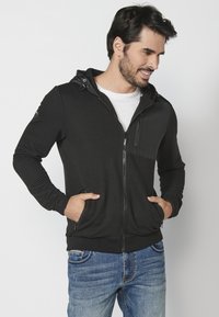 Felpa nera con zip e texture a costine, presenta un pannello frontale solido, cappuccio regolabile, tasche laterali e banda inferiore. Abbinata a jeans blu.