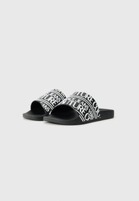 Des sandales noires à semelle plate avec de larges sangles à motifs comportant du texte blanc. La semelle est texturée pour garantir une bonne adhérence. Le design est simple et fonctionnel.