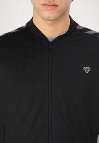 Zwarte zip-up jas met een gestructureerd oppervlak met een subtiel patroon. Grijze accenten op de schouders. Klein embleem op de linkerborst.