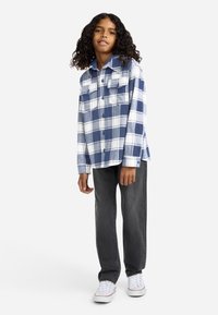 Μπλε και λευκό καρό πουκάμισο φλατέν με δύο τσέπες στο στήθος, συνδυασμένο με σκούρο γκρι τζιν και λευκά αθλητικά παπούτσια. Ορατά σγουρά μαλλιά.