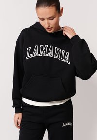 Czarna bluza z kapturem z białym napisem "LAMANIA", kieszenią z przodu i ściągaczami. W połączeniu z czarnymi spodniami dresowymi z haftowanym logo. Gładki materiał.