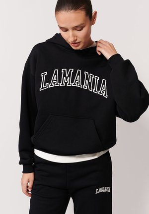 Czarna bluza z kapturem z białym napisem "LAMANIA", kieszenią z przodu i ściągaczami. W połączeniu z czarnymi spodniami dresowymi z haftowanym logo. Gładki materiał.