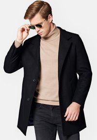 Czarny płaszcz z mieszanki wełny o dopasowanym kroju i klapach z wycięciem, nałożony na beżowy sweter z golfem. Posiada dwa guziki i kieszenie.