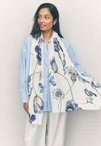 Écharpe florale présentant des motifs bleus et verts sur fond blanc. Tissu léger avec bords à franges. Portée drapée sur une chemise bleu clair.