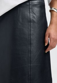 Jupe en cuir noir à la texture lisse, avec une couture latérale et une légère brillance. Main visible effleurant doucement le tissu.