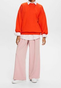 Sudadera de gran tamaño de color naranja sobre una camisa blanca y rosa claro a rayas, combinada con pantalones de pierna ancha rosa claro y zapatillas blancas.