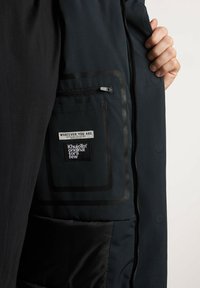 Cappotto nero con finitura testurizzata, dotato di chiusura con zip, tasca interna con etichette e fodera liscia.