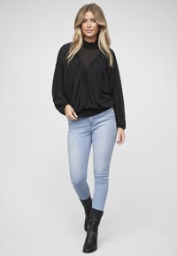 Schwarze Chiffonbluse mit hohem, gerafftem Kragen und weiten Ärmeln, kombiniert mit hellblauen Skinny Jeans und schwarzen Stiefeletten.