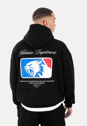Mand iført sort hættetrøje med teksten "Athleisure Department" og blå-rødt rullende katte-logo på ryggen, stående med ryggen mod på hvid baggrund.
