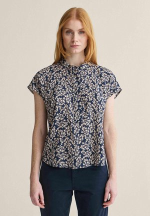 Kvinde med rødt hår iført en marineblå kortærmet bluse med beige blomsterprint og mørke bukser, stående foran en ensfarvet baggrund.