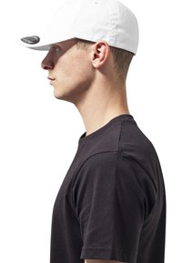 Hvid kasket med en buet skygge, der viser et mørkt logo på siden; båret med en sort kortærmet t-shirt, vist i profilvis.