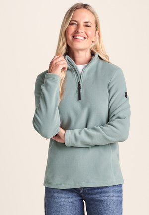 Smiling woman wearing a light green fleece pullover with a half-zip collar and blue jeans, posing with one hand adjusting the zipper.