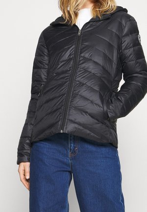 Kvinde iført en sort quiltet jakke med lynlås og hætte samt blå jeans, stående med den ene hånd i lommen mod en lys baggrund.