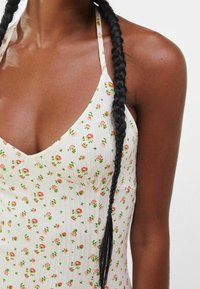 Mörkhyad person med långt flätat hår som bär en vit halterneckklänning med små röda och gröna blommönster.