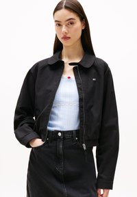 Sort kort jakke med krave og lynlås foran, med en lille logo-patch. Båret over en lyseblå top og mørke jeans. Glat tekstur.