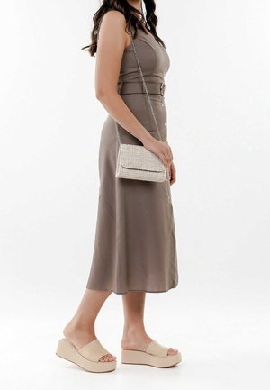 Frau steht, trägt ein ärmelloses taupefarbenes Midikleid, beige Plateausandalen zum Hineinschlüpfen und hält eine kleine beige texturierte Umhängetasche.