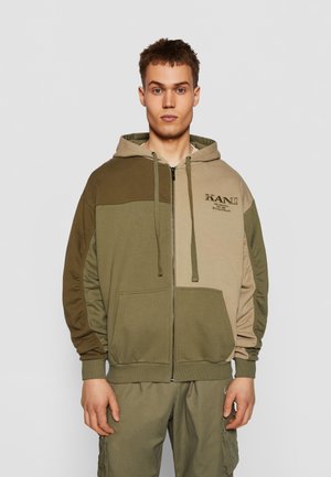 Zippertrøje i olivengrøn og beige. Har snoretræk, en kengurulomme og et broderet logo. Blødt, struktureret stof.