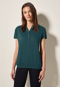 Polo top verde scuro a maniche corte con scollatura a V e placca con tre bottoni; presenta un orlo arricciato e un tessuto morbido e leggero.