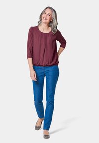 Bordeaux blouse met een geplooide voorkant en driekwart mouwen, gecombineerd met blauwe jeans en lichtgrijze ballerina's met een strikaccent.