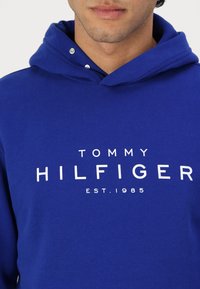 Blaue Kapuzenjacke aus Baumwolle mit einem Frontlogo in Weiß, das "TOMMY HILFIGER EST. 1985" liest. Das Design umfasst eine Kapuze mit Kordelzug.