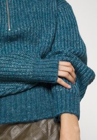Pull en tricot teal avec des poignets côtelés et une texture ; possède un col zippé et une coupe décontractée, associé à une surface en cuir brun matelassée.