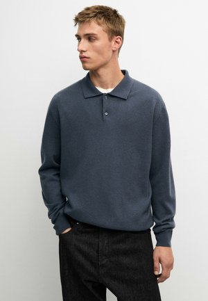 Junger Mann mit kurzen Haaren, der ein dunkelblaues Langarm-Poloshirt und schwarze Hose trägt, schaut nach links mit den Händen in den Taschen.