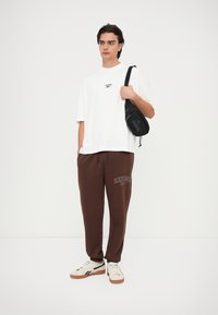 Camiseta blanca de manga corta con logo impreso, pantalones deportivos marrones con texto "Reebok" y zapatillas beige con acentos negros. Bolsa bandolera en negro.