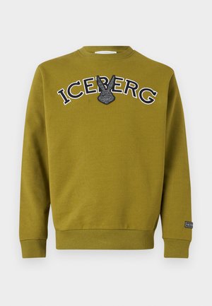 Junger Mann trägt olivgrünes "ICEBERG" Sweatshirt, schwarze weite Hose, schwarze Kappe und grüne Sneakers mit roten Schnürsenkeln, steht vor einfarbigem Hintergrund.