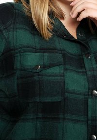 Zielono-czarna flanelowa koszula w kratę z metalowymi zatrzaskami i dwoma kieszeniami na piersiach. Materiał ma miękką fakturę i utrzymaną strukturę.