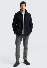 Pardessus in lana blu navy sopra una giacca nera con zip, una maglietta bianca, pantaloni grigi e stivali neri alla caviglia, con bottoni doppi e una finitura texturizzata.
