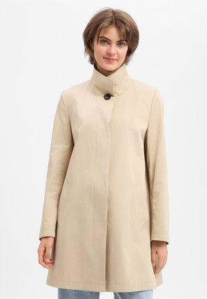 Frau trägt einen beige-farbenen kniehohen Mantel mit hohem Kragen und einem einzelnen schwarzen Knopf, steht vor einem schlichten weißen Hintergrund.