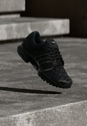 Zapatilla deportiva negra con un patrón texturizado, sistema de cordones y suela acolchada. Parece flotar sobre una superficie de concreto.