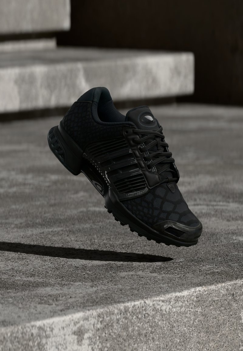 Must kergejõustikujalanõu tekstuurse mustriga, paeltega ja pehmendusega tallaga. Tundub, et see hõljub betoonpinna kohal.