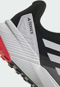 Zapatilla deportiva negra y gris con parte superior de malla texturizada, acentos blancos, suela de goma con detalles rojos y la marca "TERREX".