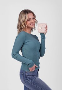 Teal ribbed long-sleeve top paired with blue jeans. Model holds a light pink wallet with gold accents. Soft texture, fitted design.