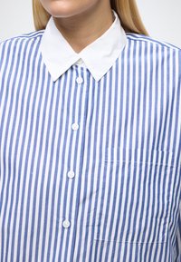Camicia a righe blu e bianche con colletto bianco, dotata di chiusura con bottoni e una piccola tasca sul petto. Realizzata in tessuto liscio.