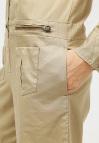 Tuta utilitaria beige con tasca con zip e due tasche frontali a toppa. Tessuto strutturato con un fit rilassato e un design semplice e funzionale.