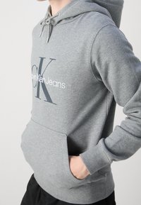 Persona vistiendo una sudadera gris de Calvin Klein Jeans con las manos en el bolsillo frontal, de pie contra un fondo claro y liso.